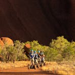 A group of Segway riders appear tiny against the massive formation that is Uluru, towering over them