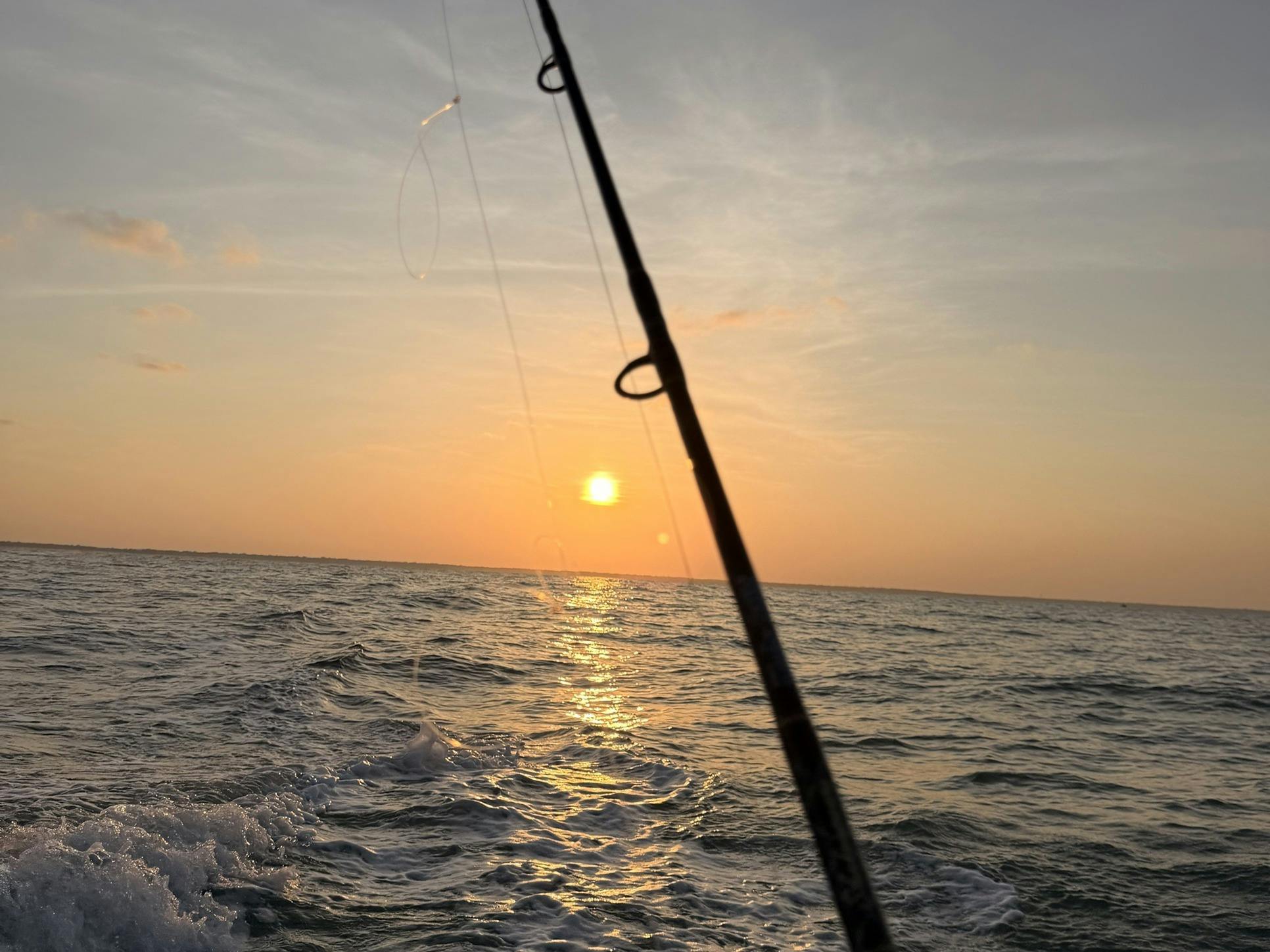 A fishing rod angled across a golden sunset, with the boat's wake on the open sea below.
