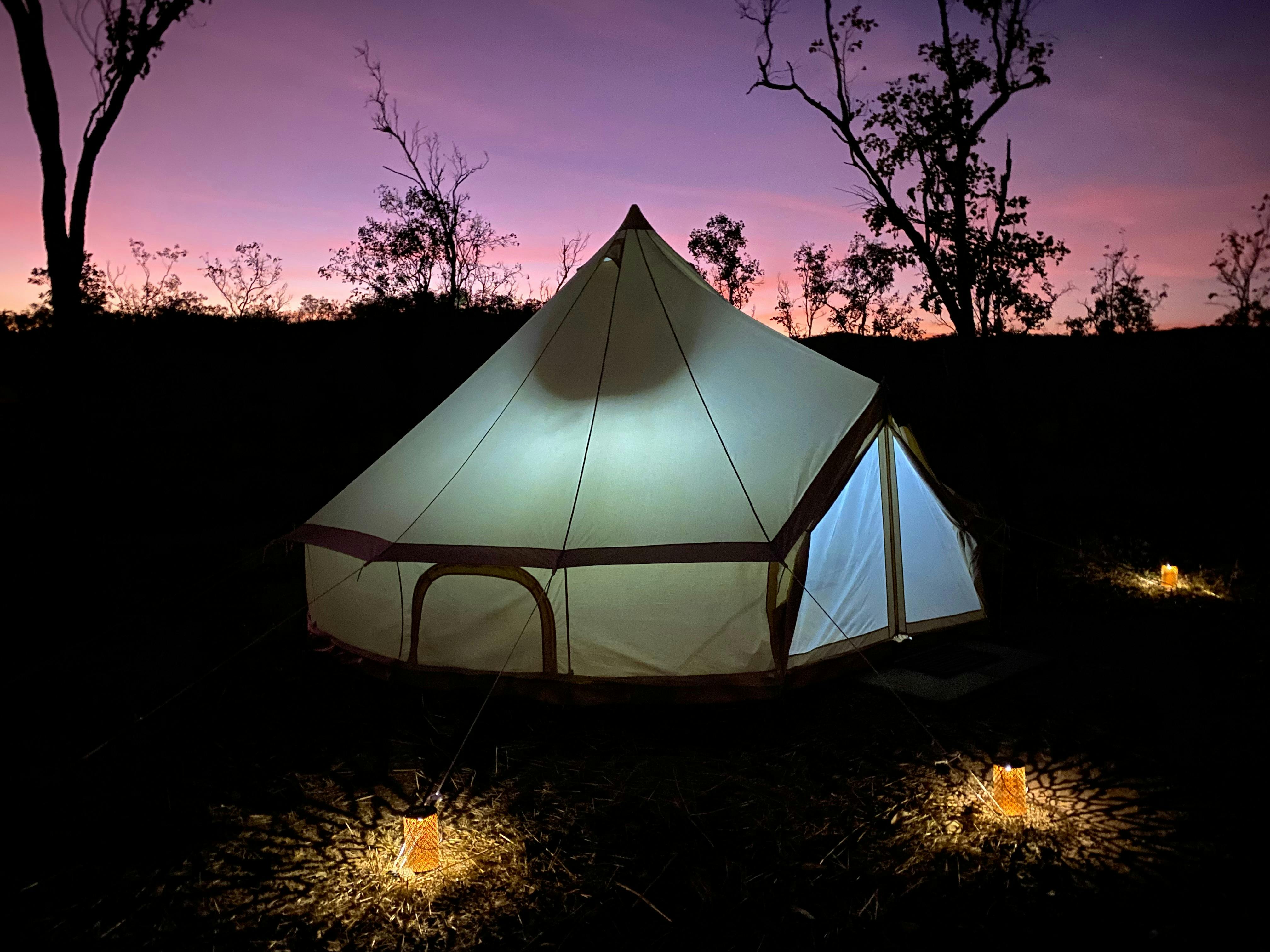 A glamping canvas bell tent sits against a purple evening sky surrounded by solar lanterns