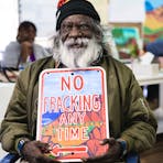 An Aboriginal artist sits holding an artwork which reads "nom fracking any time"