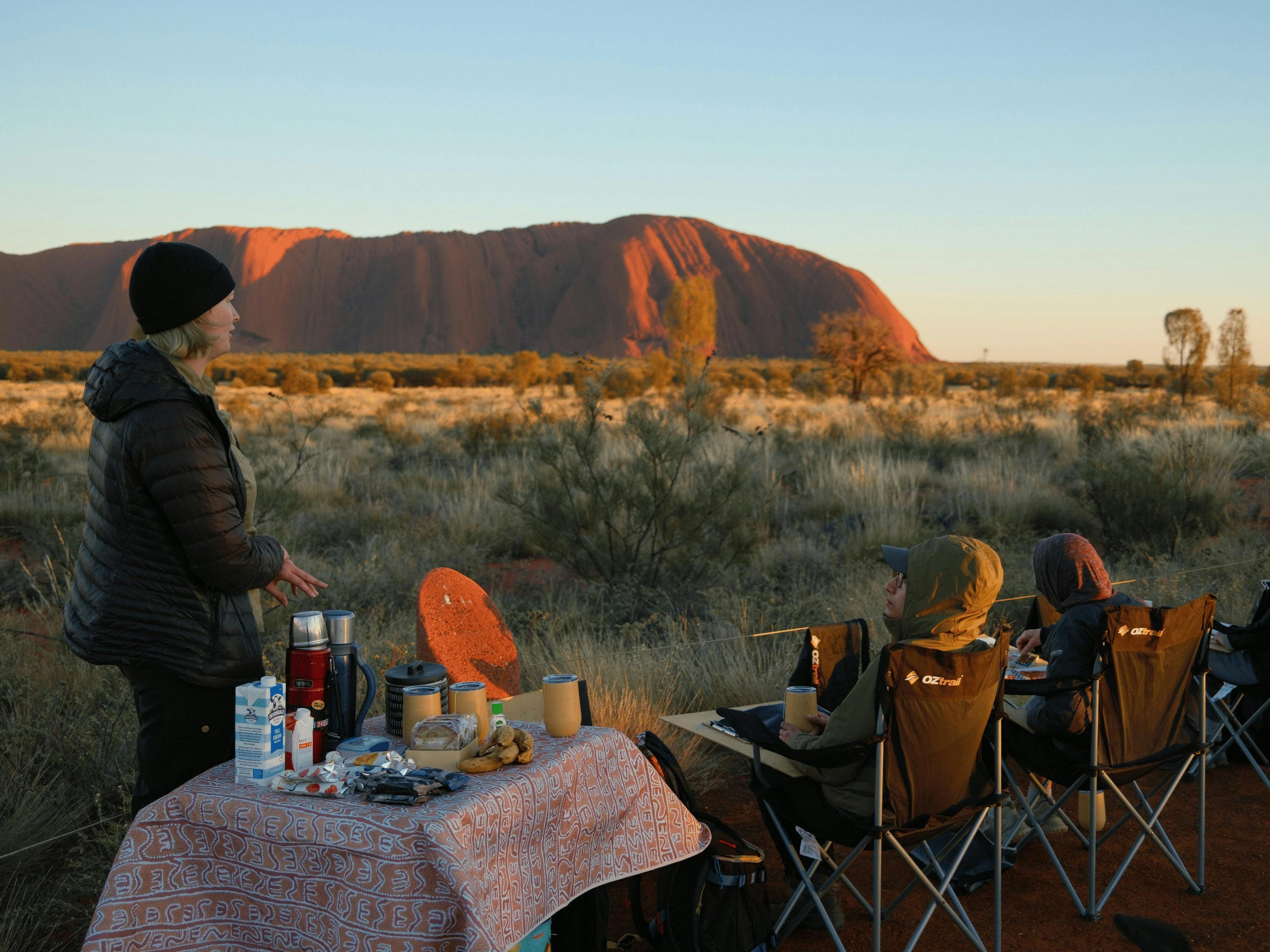 A woman stands at a breakfast table outdoors talking to two people sitting with Uluru behind