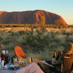 A woman stands at a breakfast table outdoors talking to two people sitting with Uluru behind