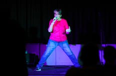 Woamn onnstage wearing jeans and pink teeshirt speaking into microphone