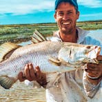 Man standing in shallow, muddy floodplain water holding a large barramundi horizontally.