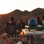 Camping stove and billy with campers in the background sitting around a fire looking at the sunset