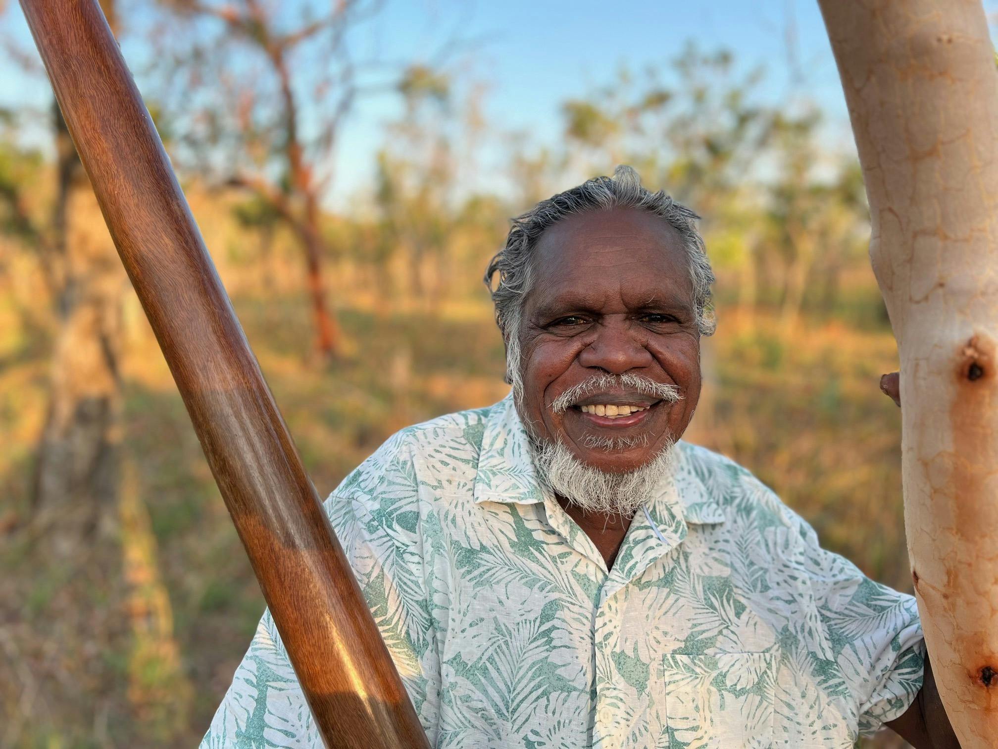 Aboriginal man Manuel Pamkal stands next to a tree with didjeridoo