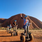 A group of Segway riders zip past the camera with a brilliant blue sky and red Uluru behind them.