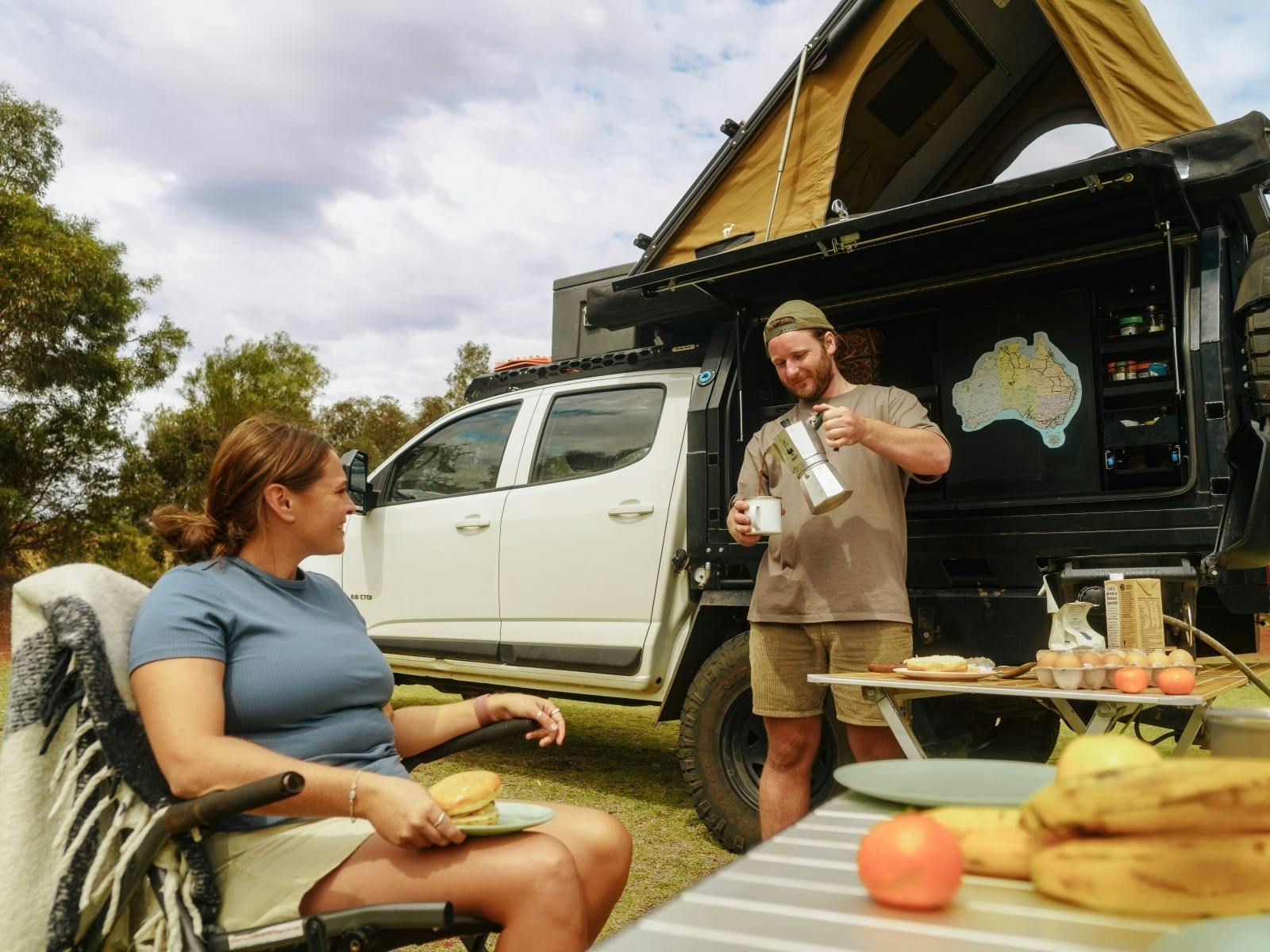 A person standing and pouring coffee into a mug in front of their car with person sitting in front