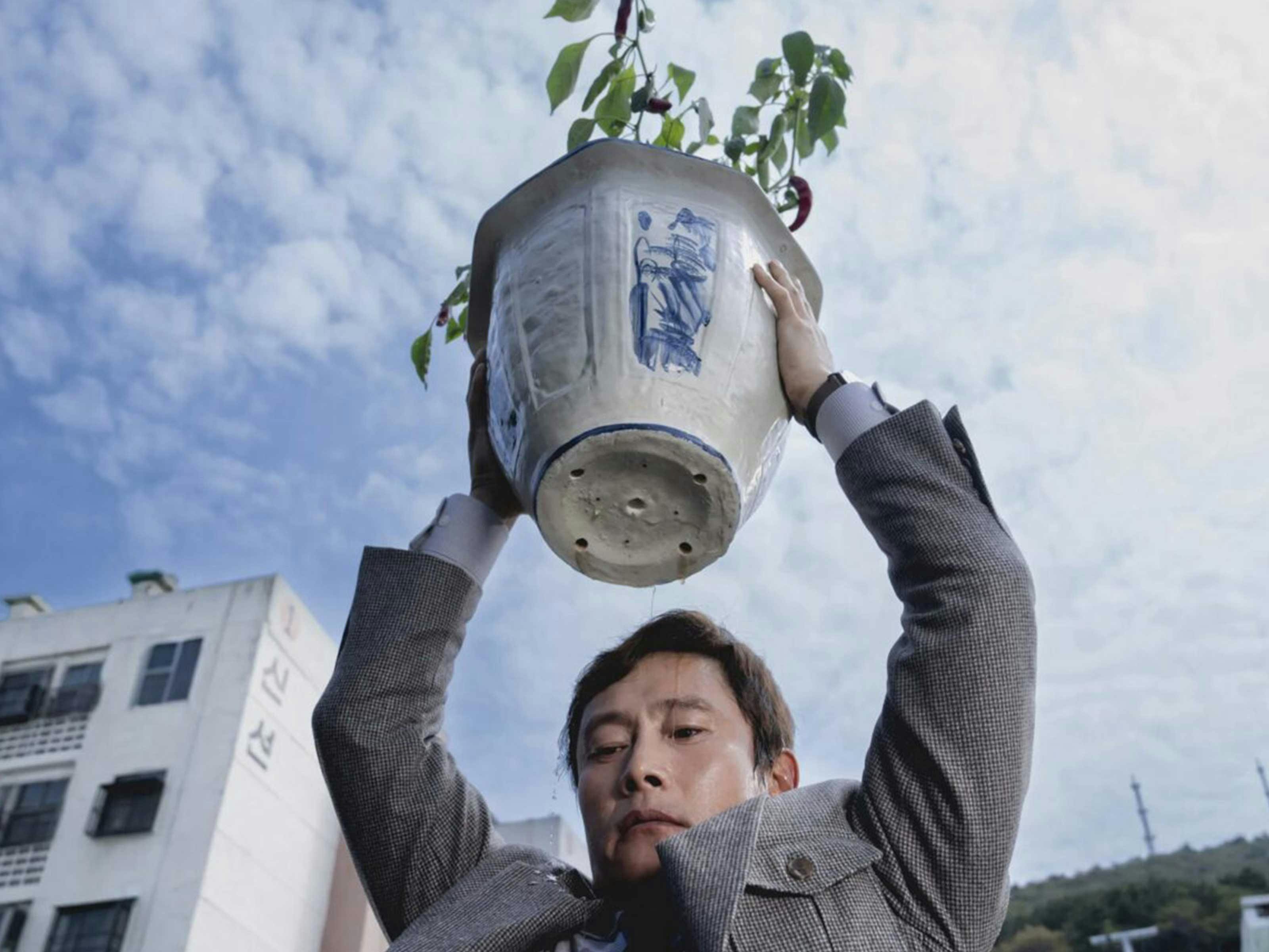 A man holds a large pot plant above his head, poised to throw it. Water drips down his face.