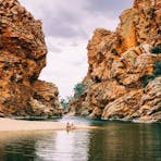 Two friends sitting on the banks of the waterhole with towering red escarpments in the background