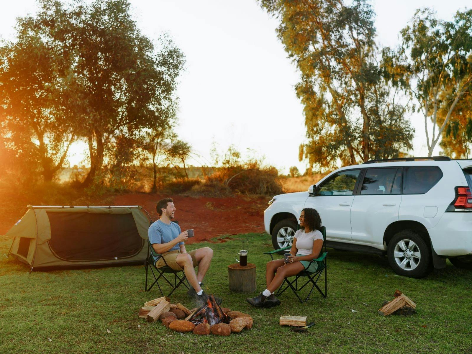 A couple sitting on camp chairs holding a cup in their hand, in front of their tent and car