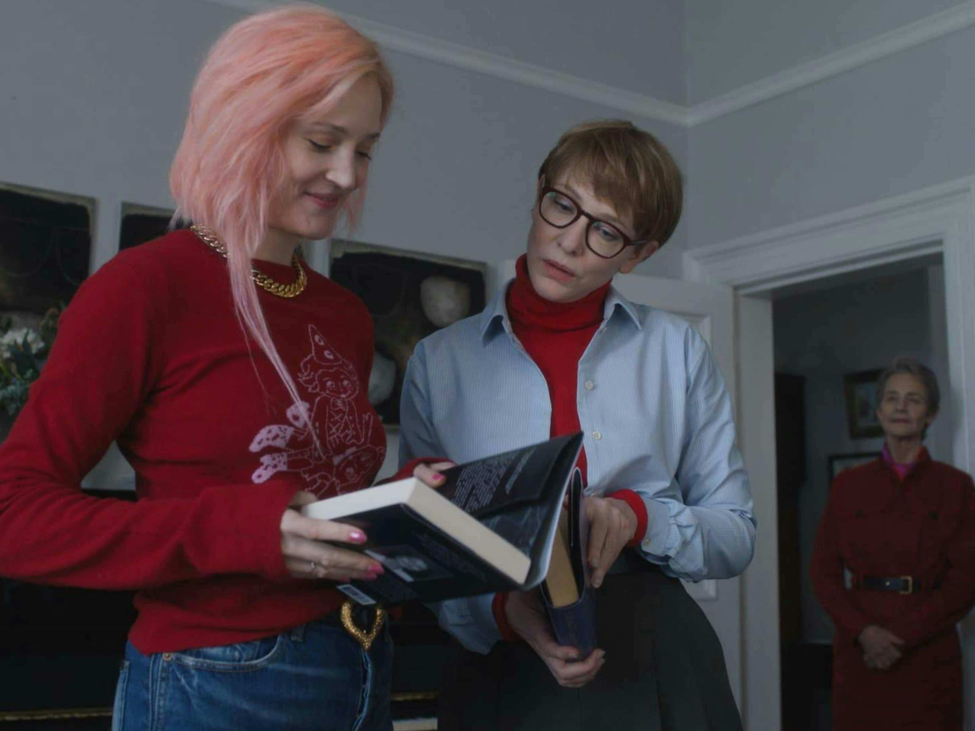 A young woman with pink hair shows an older woman with glasses a book