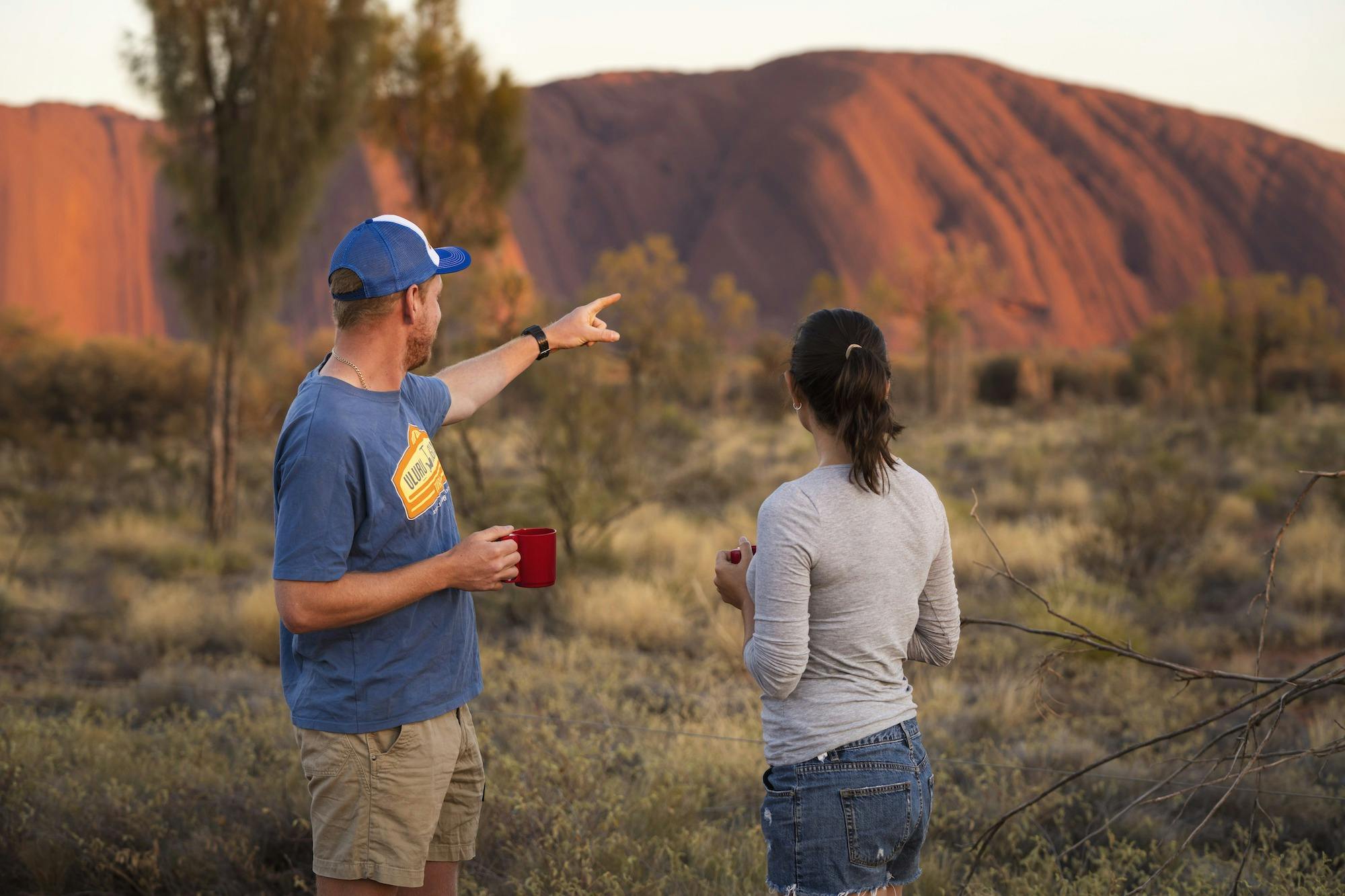 A guide points at Uluru, glowing in the morning. He shares fascinating stories with a participant.