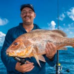 Smiling man on a fishing boat holding a large golden snapper with orange scales, with a fishing rod.