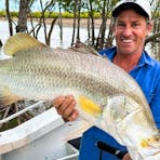 Man standing on a boat in a mangrove creek holding a large barramundi horizontally