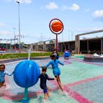 Children play in the shallow water of the splash zone and under a water fountain