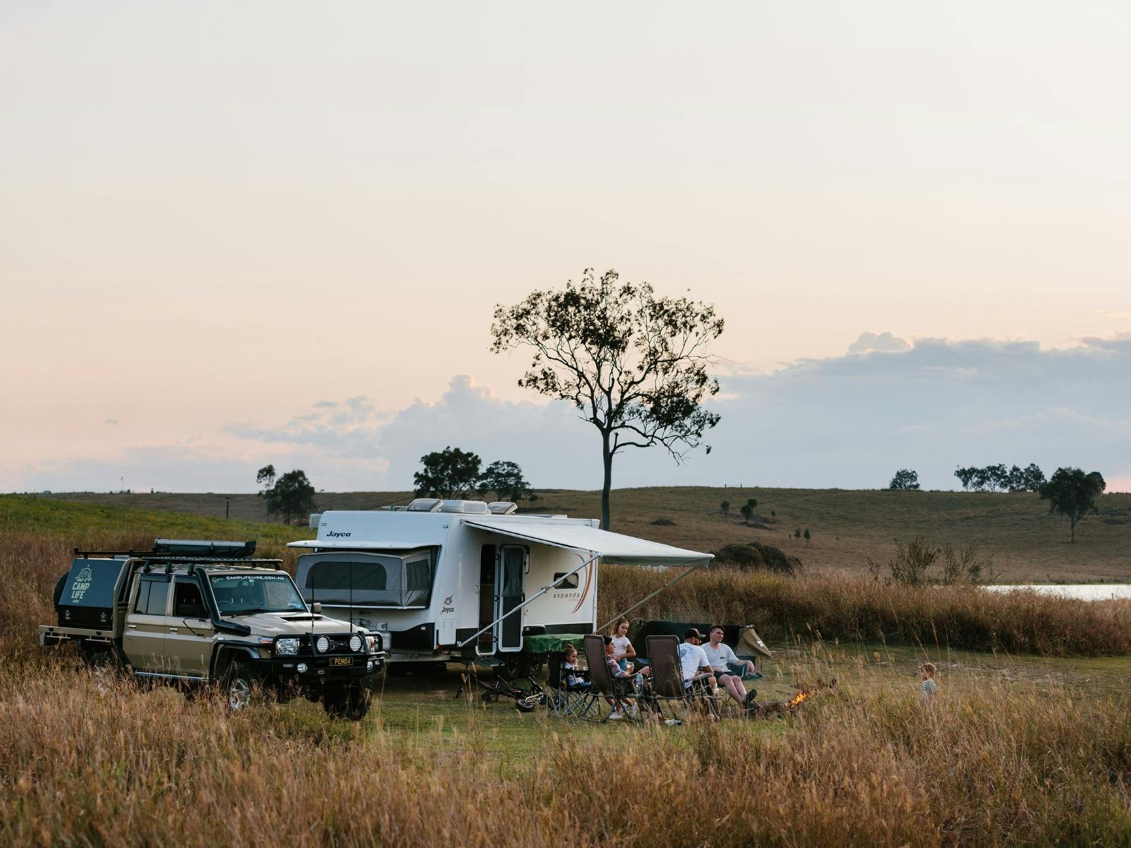 A family sits in front of their caravan, in front of a campfire