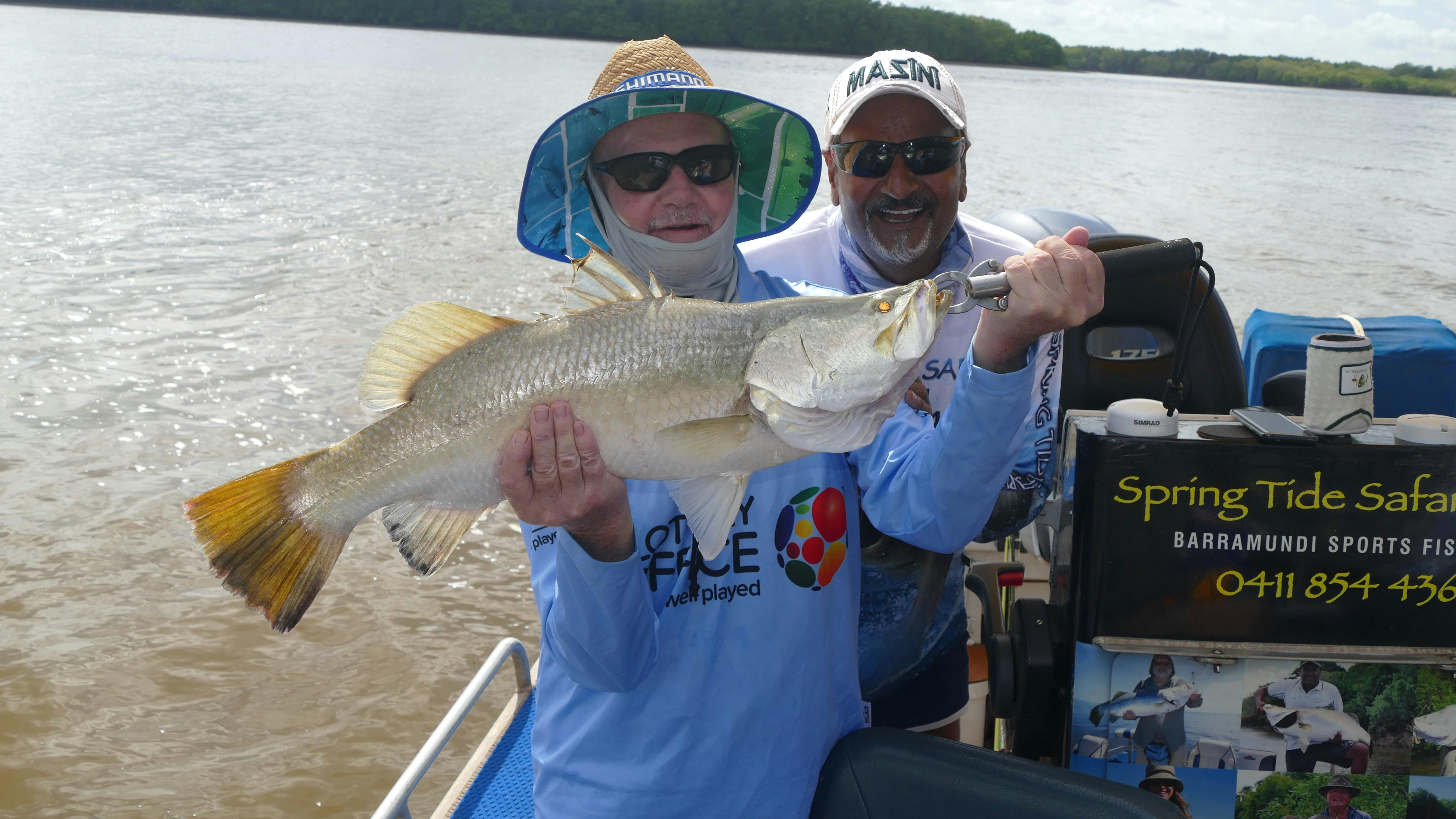 Two men standing in a boat holding a barramundi.