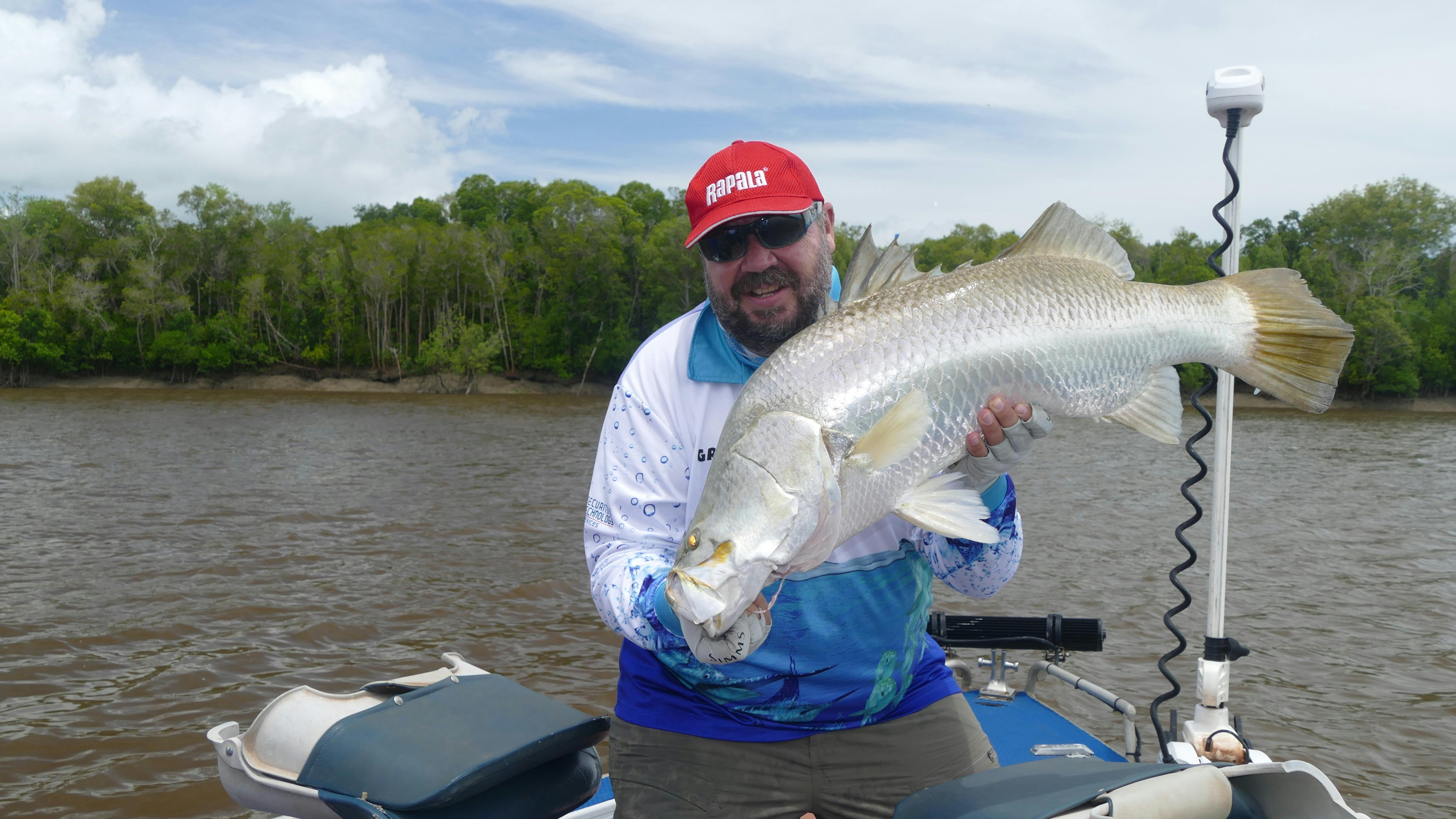 Greg Ireland standing in the boat holding a big barramundi.