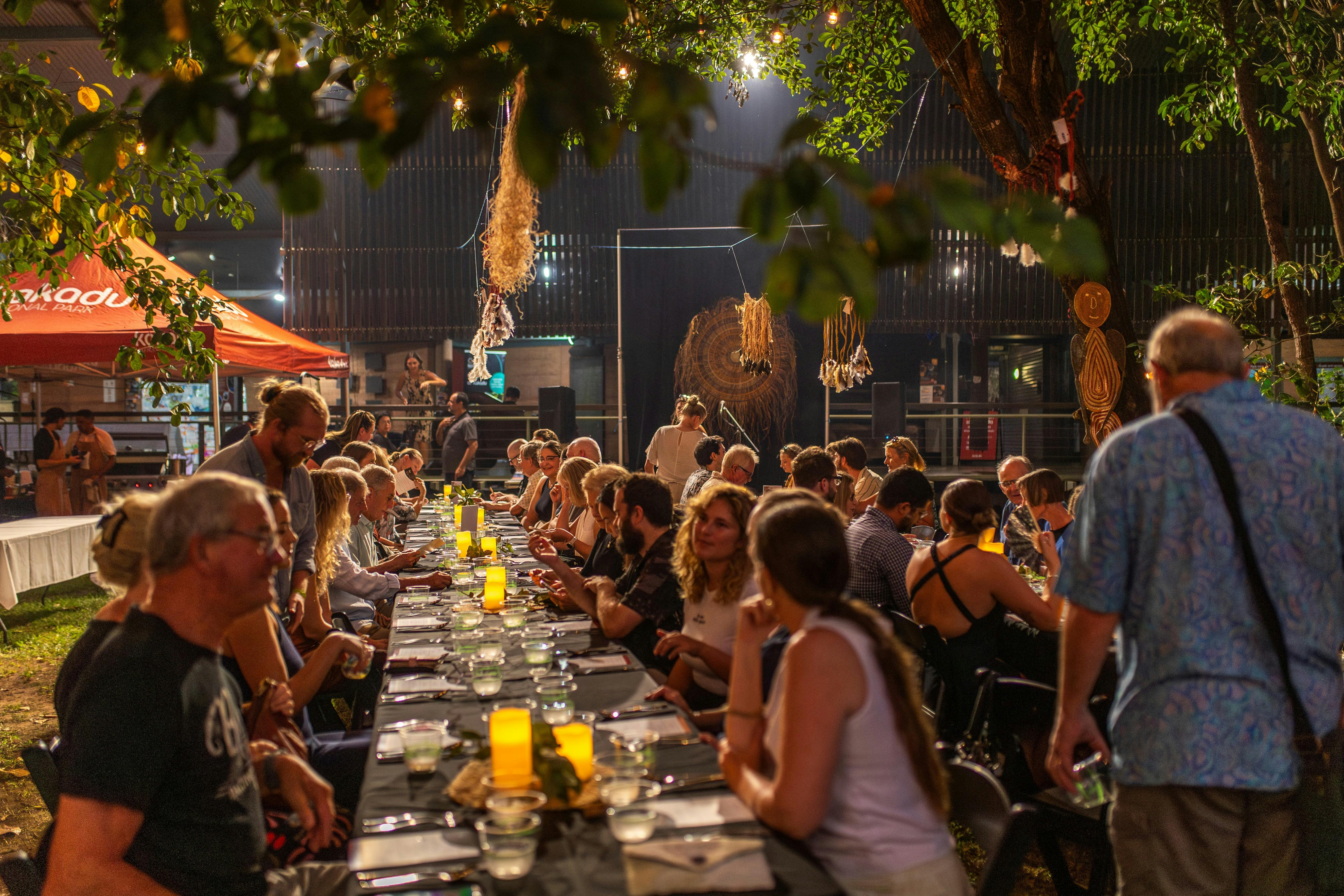 People seated at a long outdoor dinner table beneath trees and lights at night.