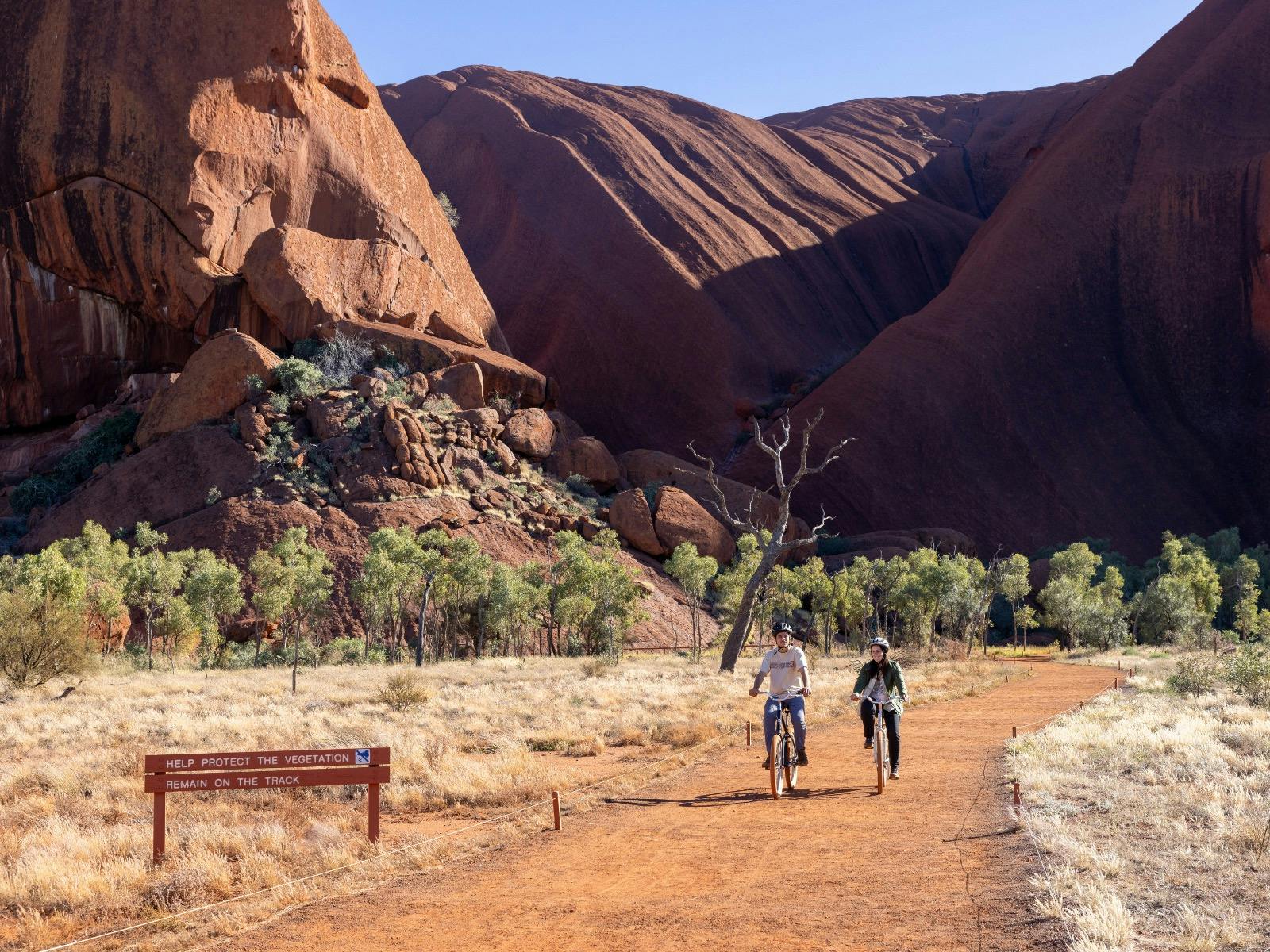 A male and female each riding a bike at the base of Uluru