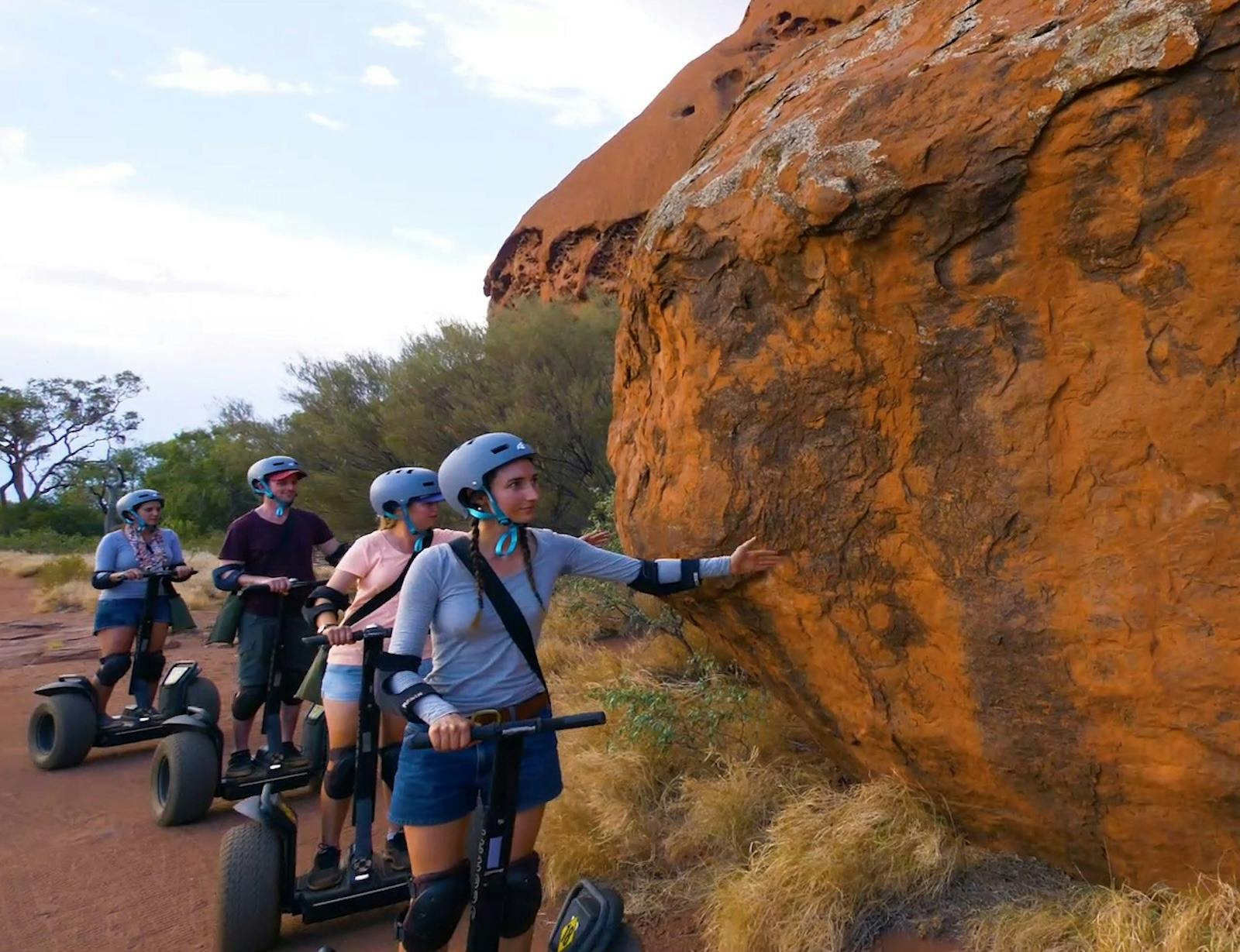 A small group of Segway riders traverses an otherwise empty path. They reach out and touch Uluru.