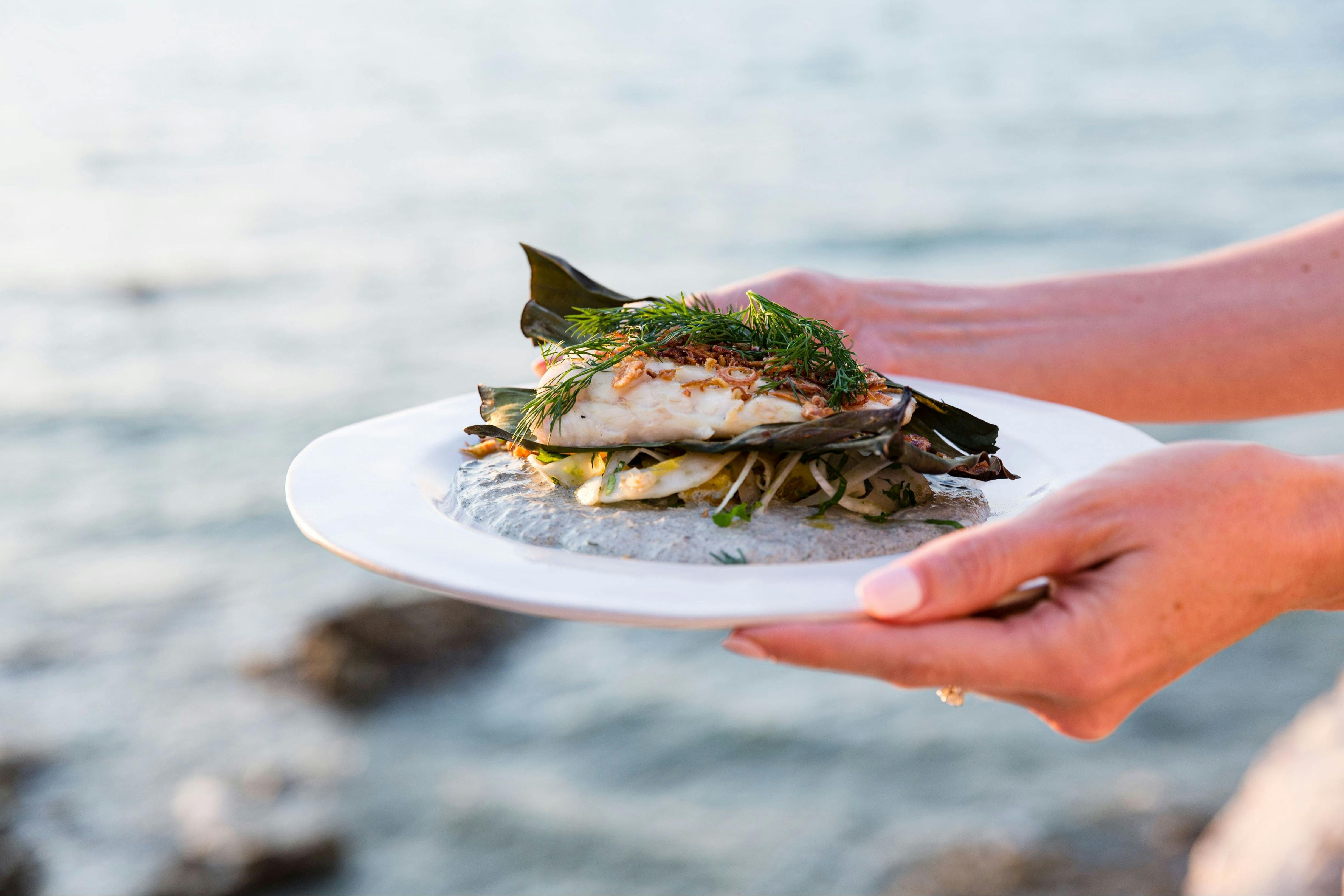Hands presenting a beautifully plated fish dish on charred banana leaves with fresh herbs.