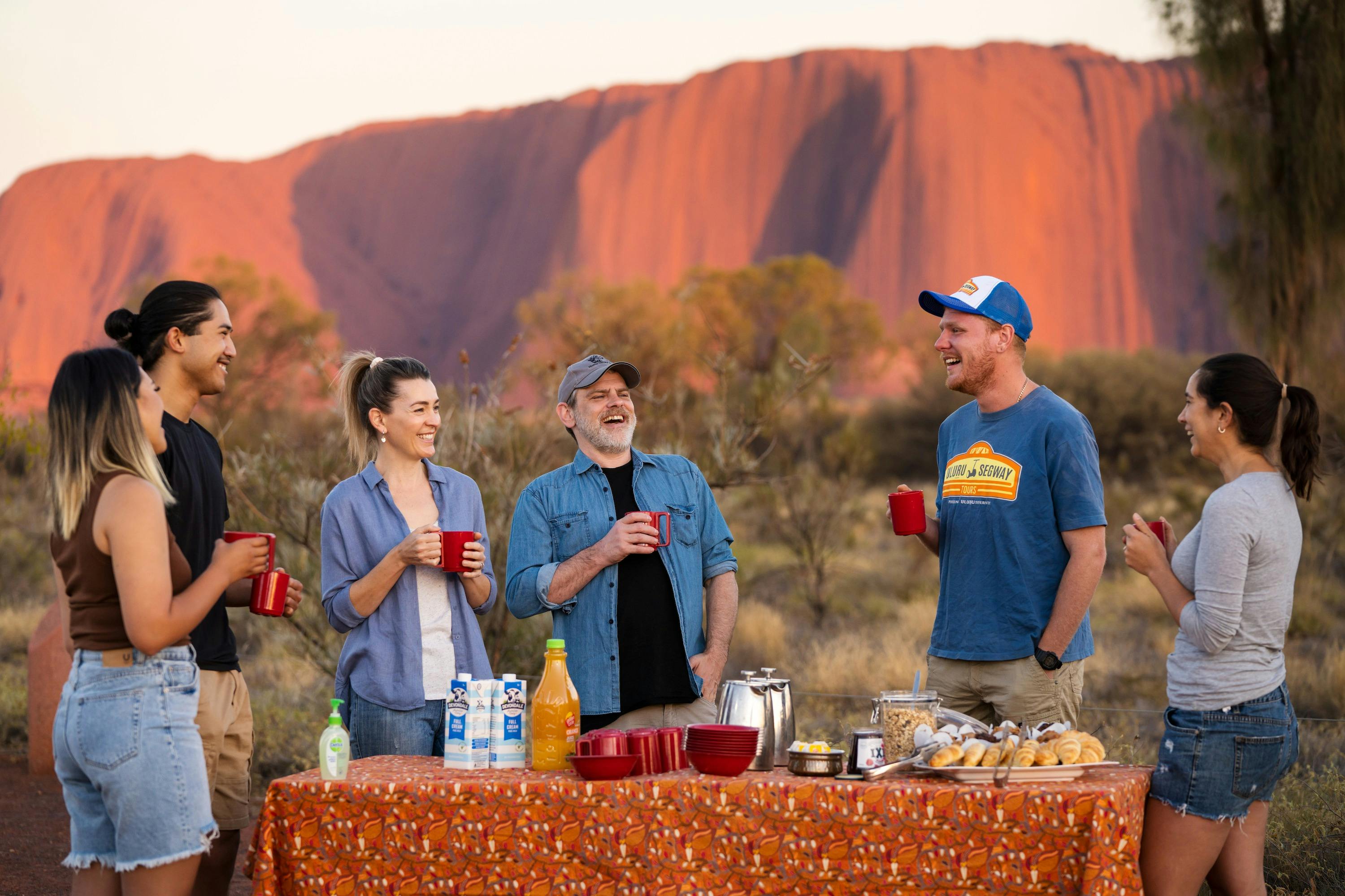 A small group of people gather around a picnic breakfast watching Uluru glow red in morning light