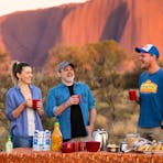 A small group of people gather around a picnic breakfast watching Uluru glow red in morning light