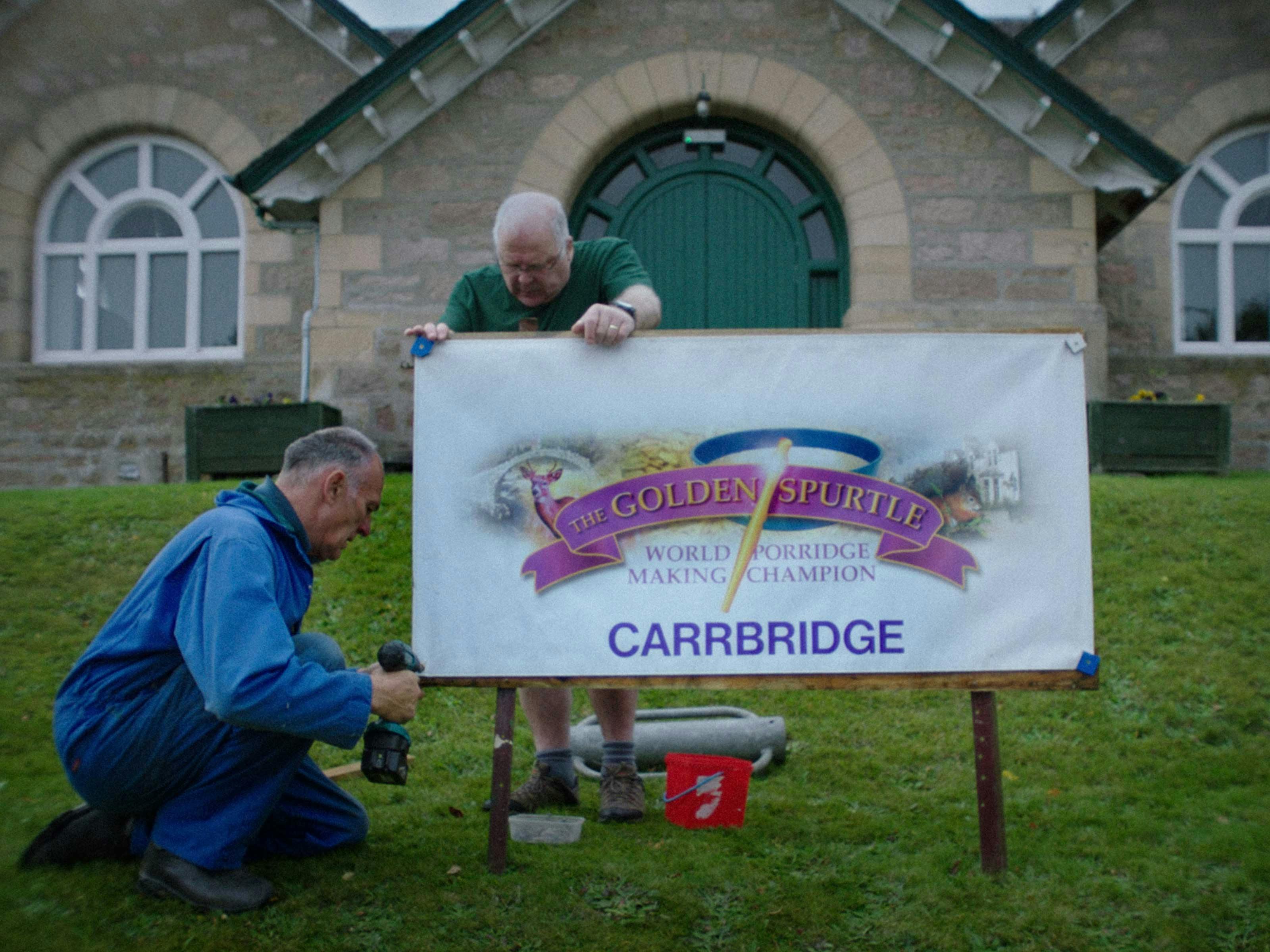 Two older men erect a sign reading "The Golden Spurtle"
