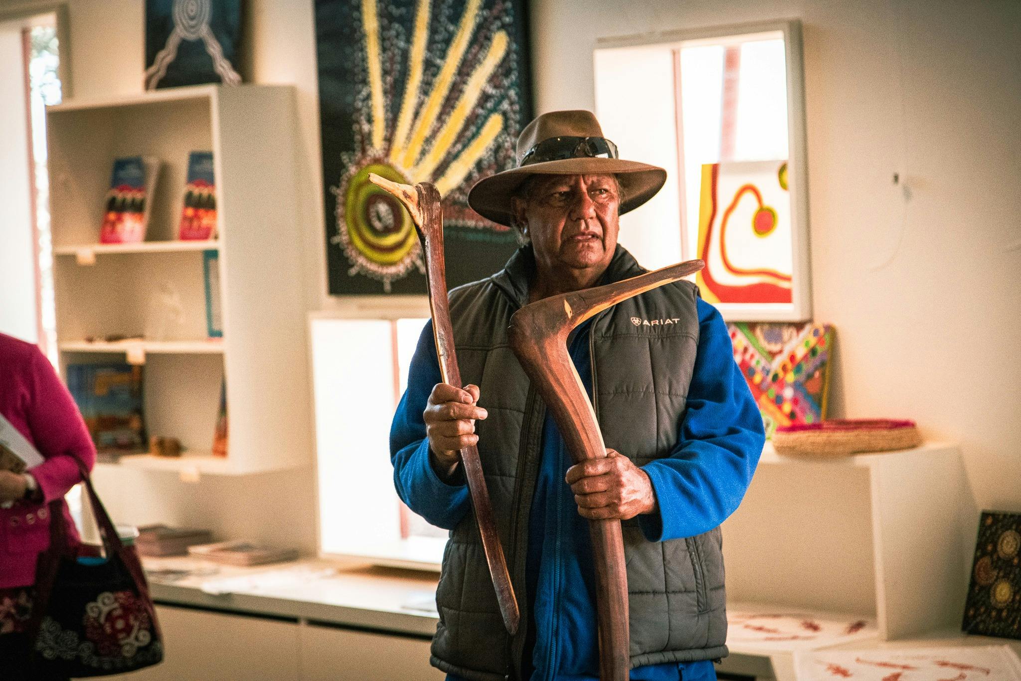 An older man with dark skin stands with a hat on holding two number 7 boomerangs inside an art store