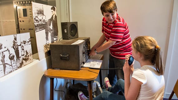 A boy operating vintage radio, girl speaking into intercom while sitting at Alice Springs School of the Air