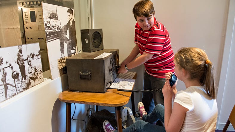 A boy operating vintage radio, girl speaking into intercom while sitting at Alice Springs School of the Air