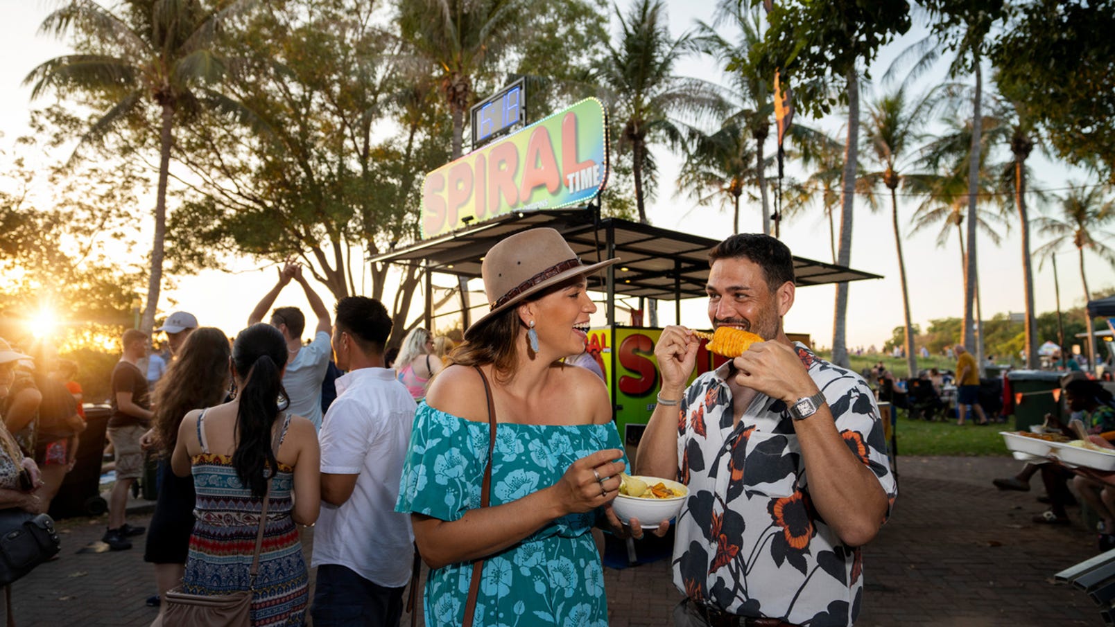 A couple enjoys laksa and potato tornado at Mindil Beach Sunset Market, bustling with people and food stalls