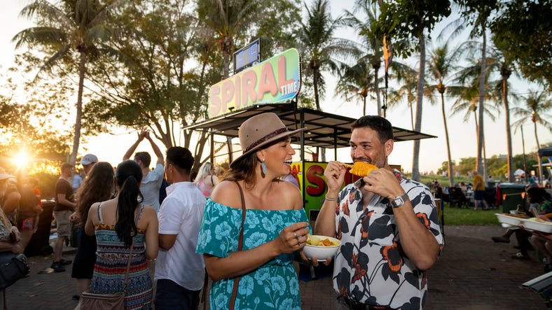 A couple enjoys laksa and potato tornado at Mindil Beach Sunset Market, bustling with people and food stalls