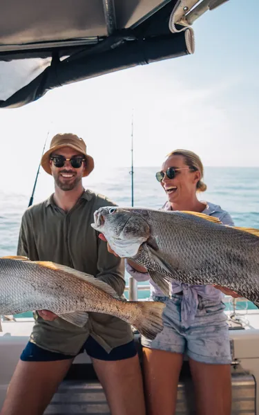 a man and a woman standing at the back of a boat with each holding a large fish