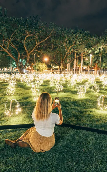 Girl sittin on the grass and using her phone to take a photo of a light installation in Darwin