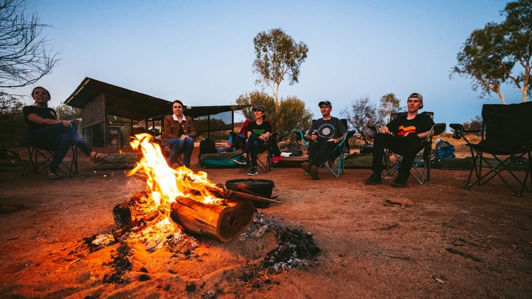 Visitors sitting around a camp fire during an overnight mountain bike adventure with Outback Cycling. Go offroad on a mountain bike tour and explore the Red Centre and it's spectacular scenery.