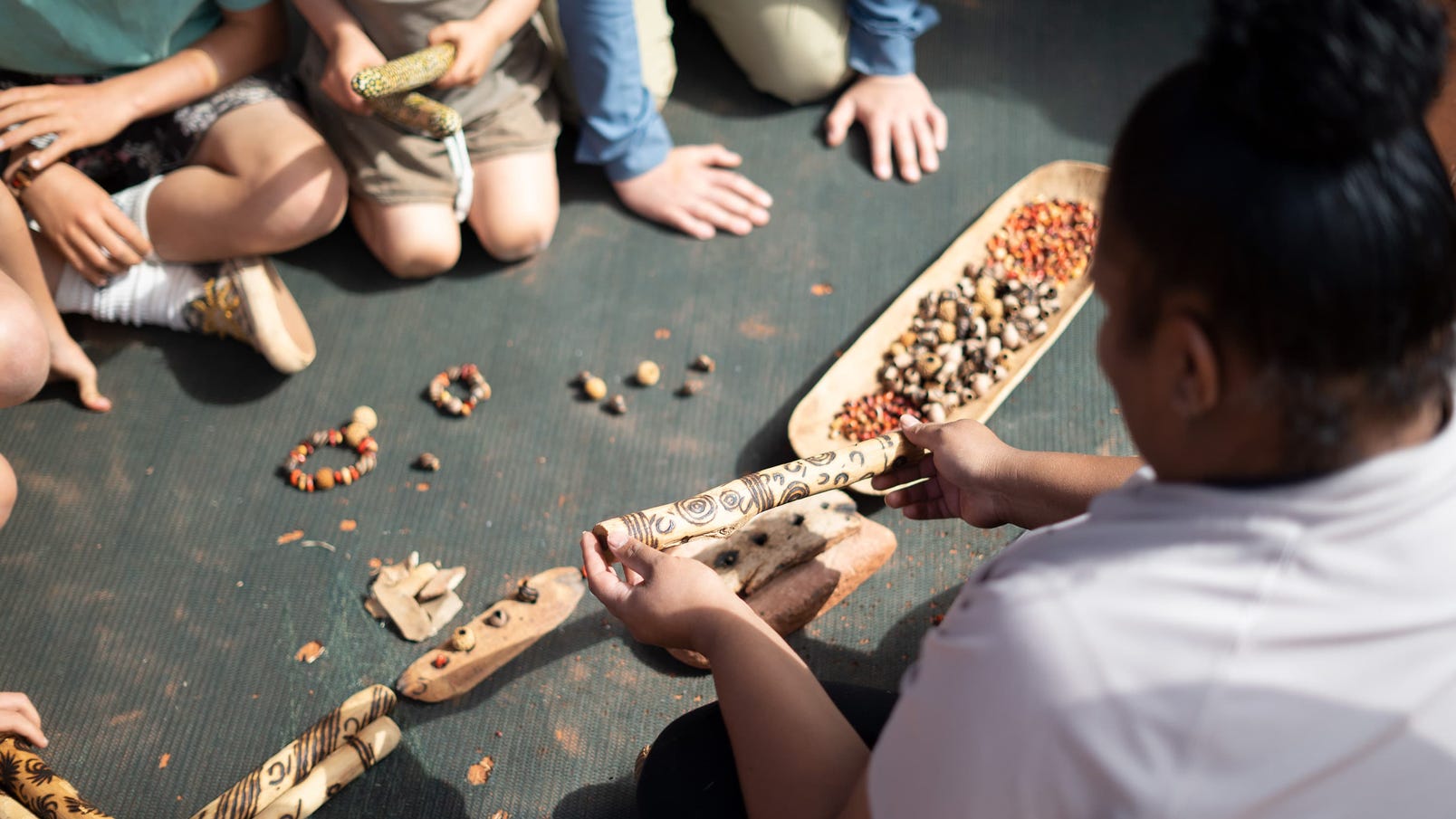 Children watching Aboriginal guide with wooden decorated artifacts, tree seeds, jewellery on floor