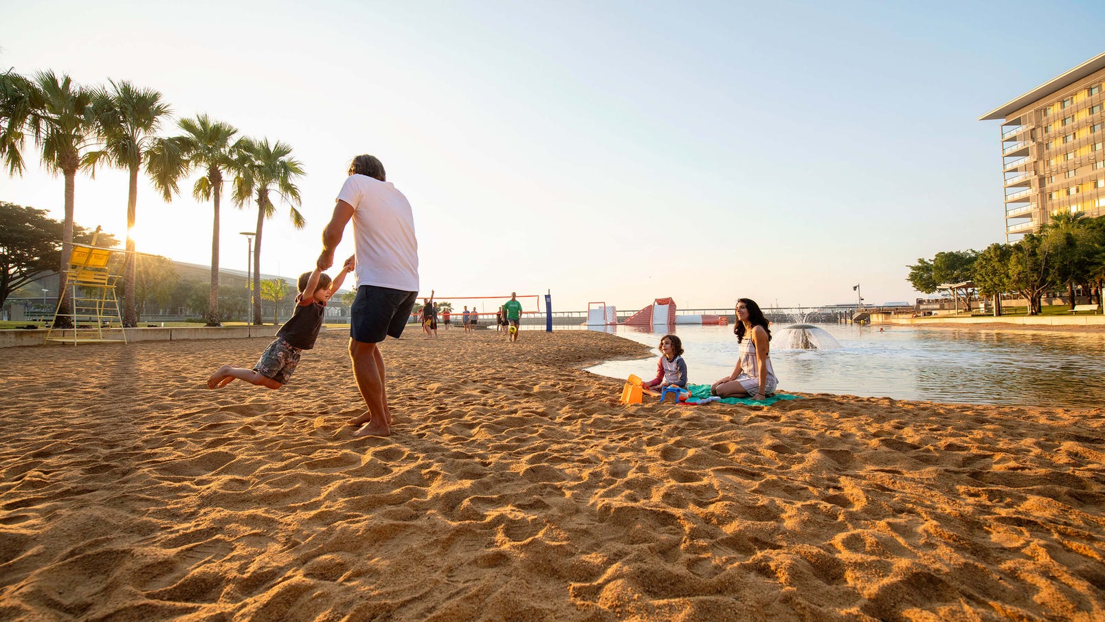 Family relaxing at the Darwin Waterfront playing beach volleyball in the background