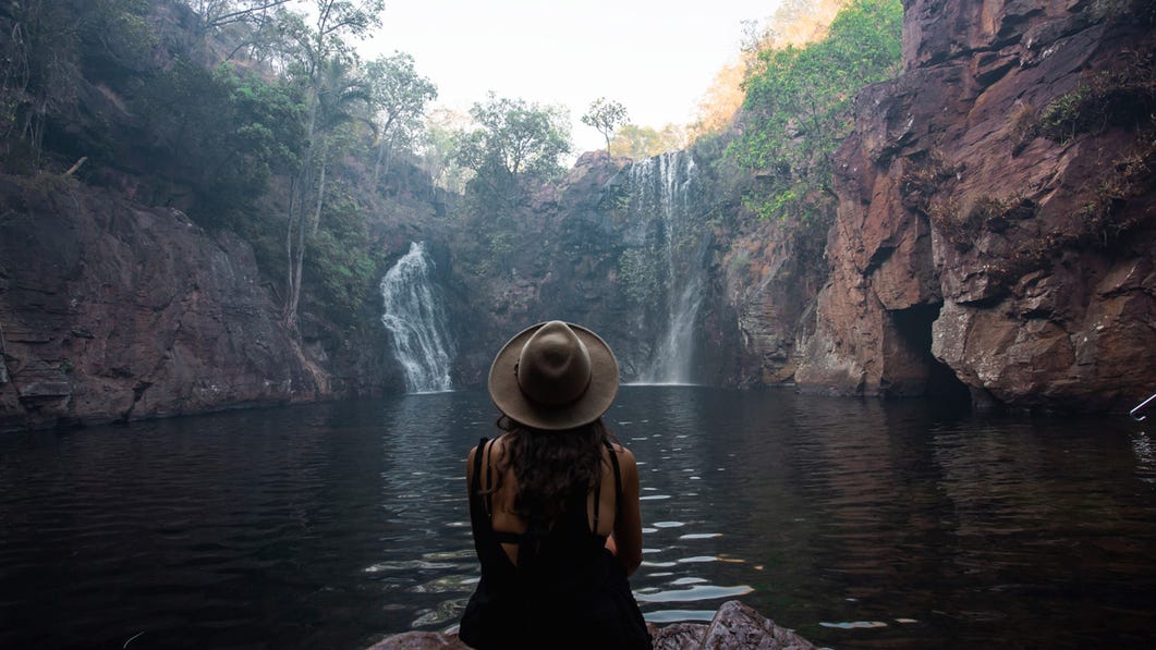 a woman sitting at the waters edge looking out at florence falls in litchfield national park