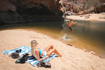 Guy jumping into the waterhole at Ormiston Gorge while his girlfriend is sunbathing on the sandy shore